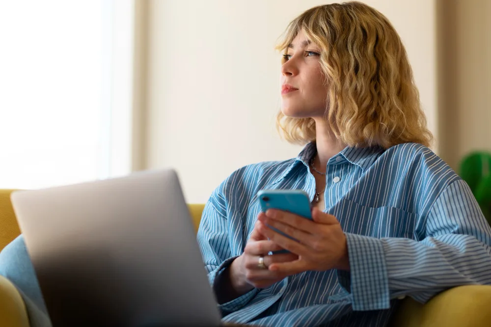 A woman on a sofa with a laptop, holding her phone and looking away