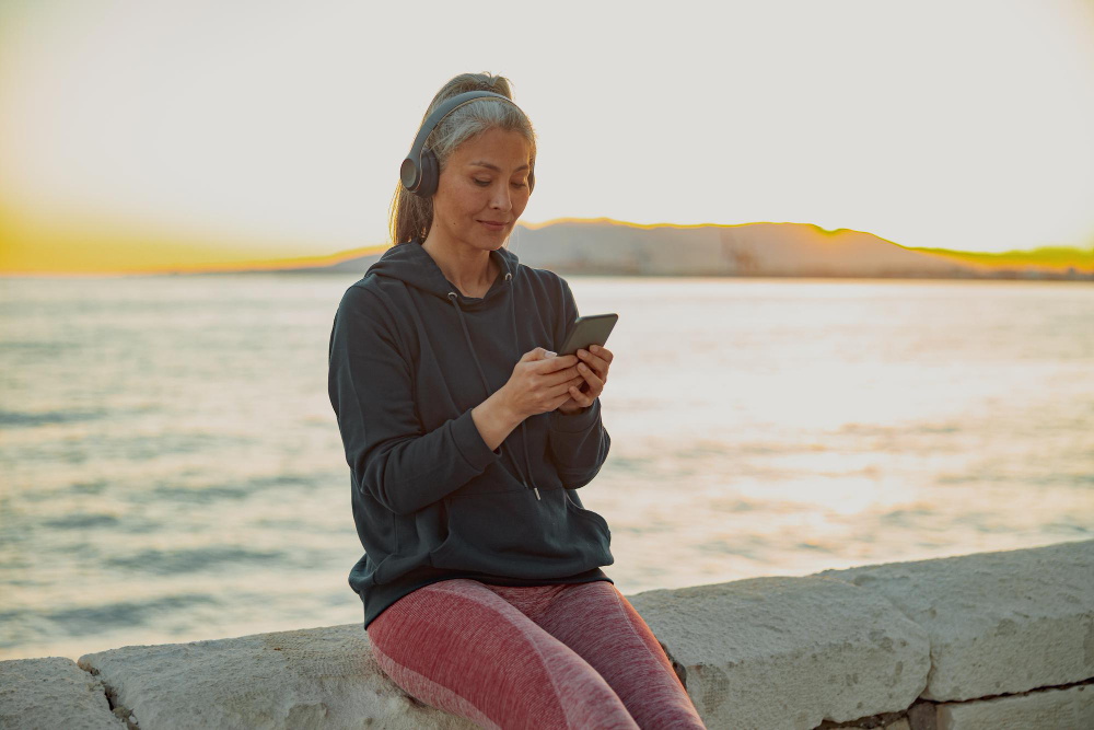 An older woman in headphones using her phone by the sea at sunset