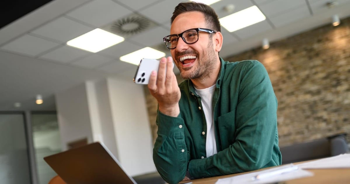A man smiling while speaking into his phone on speaker at a desk