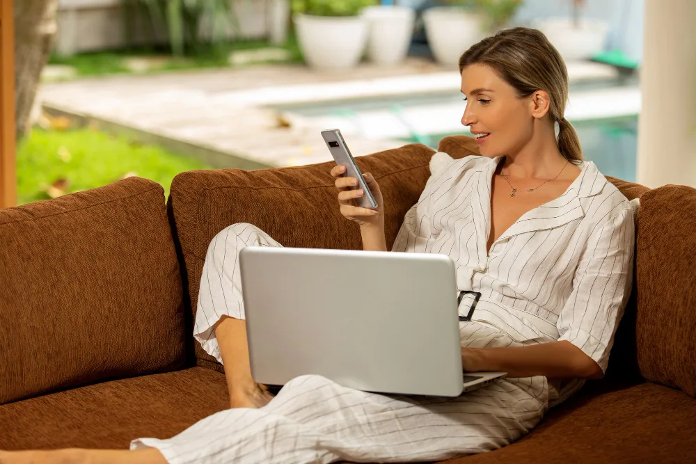 A woman on a sofa checking her phone with a laptop open