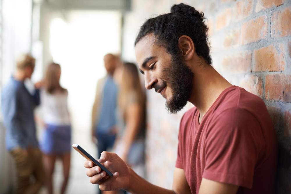 A man leaning against a brick wall, smiling at his phone