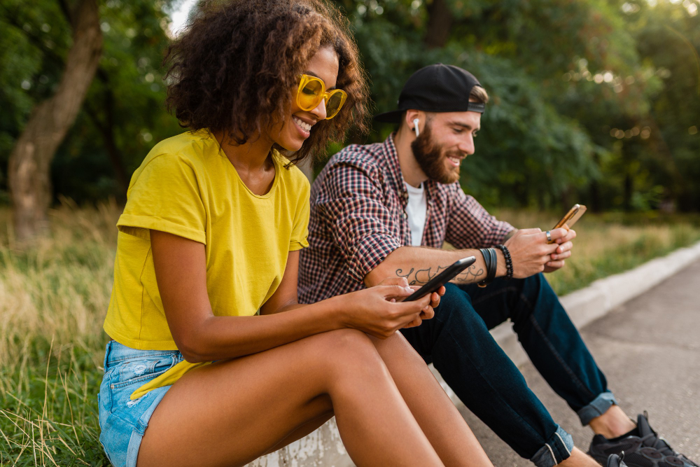 Two friends sitting on a kerb in the sun, both looking at their phones