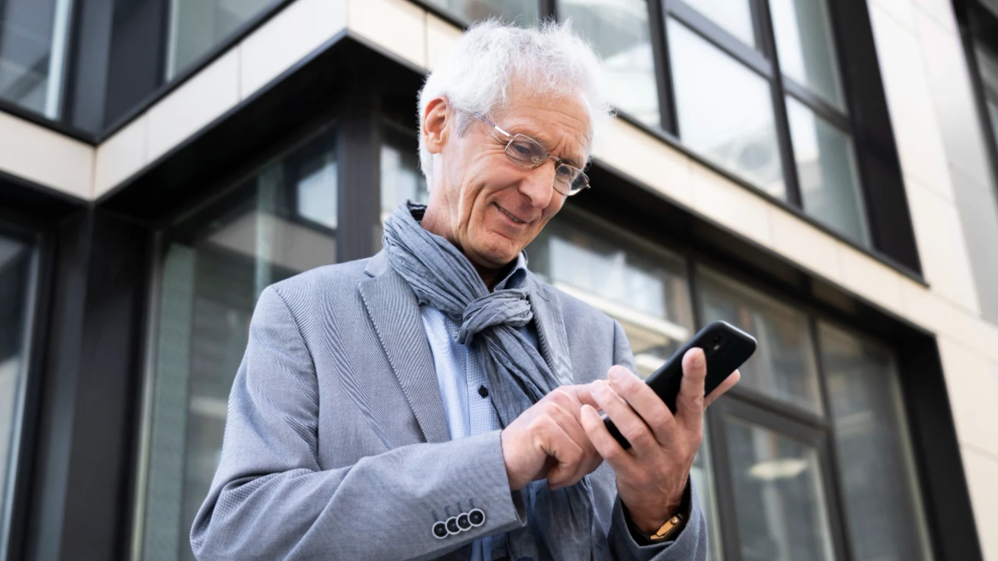 An older man in a grey jacket and scarf smiling at his phone outside a modern office building
