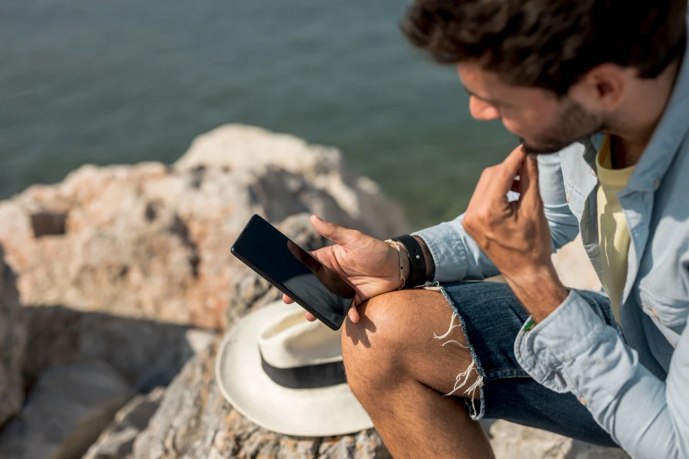 A man sitting on coastal rocks looking at his phone, open sky above the sea behind him