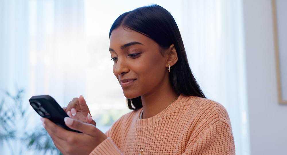 A young woman sitting by a bright window, looking at her phone