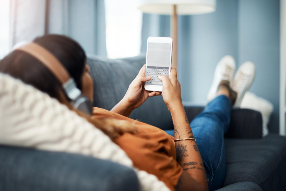 A person relaxing on a sofa in headphones, texting on a phone
