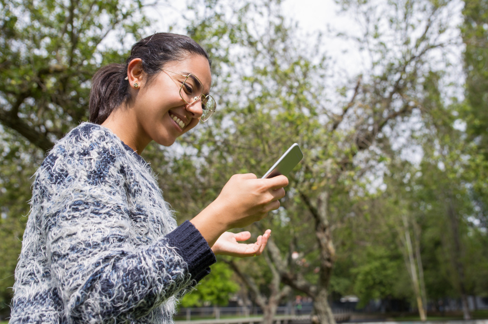 A woman in glasses smiling at her phone in a park
